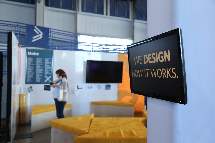 Wall-mounted monitor at The Design Lab with the text ‘We design how it works.’ A person in the background of the photo reads a poster on a wall.