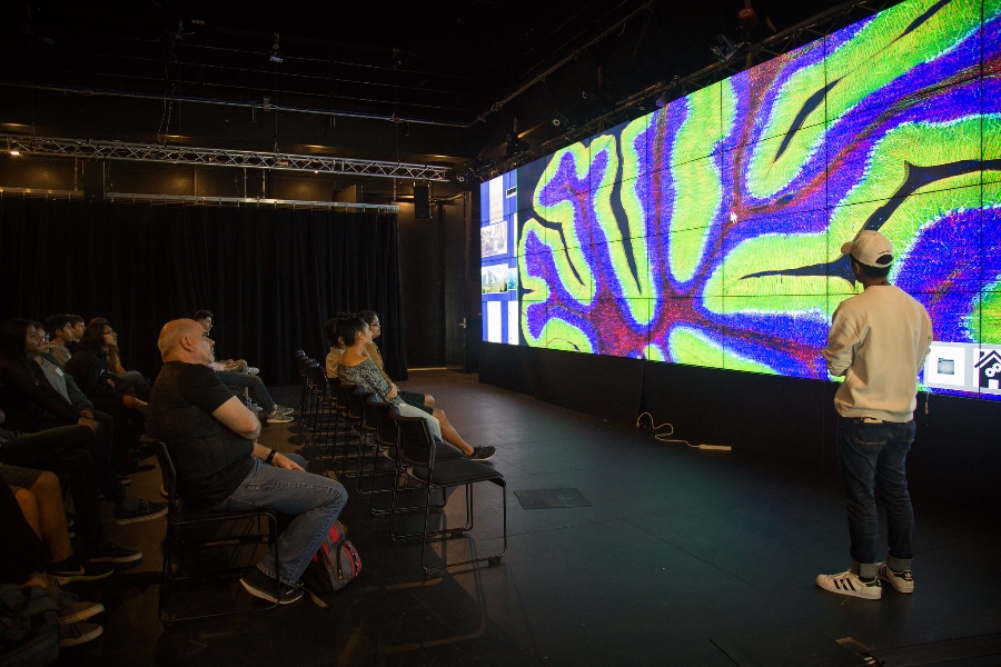 A speaker and audience at the Link2 Game Design program analyze a large LCD display of a game interface.