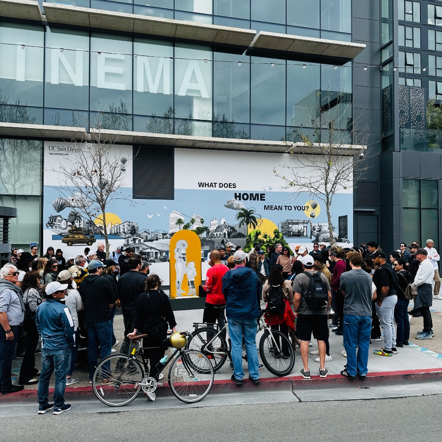 A crowd gathers around a mural displaying community icons and the text, ‘What does home mean to you?