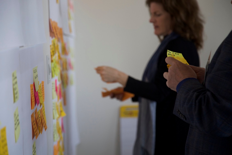 Two participants hold a stack of written post-it notes, adding them to a wall covered with other notes in a brainstorming activity